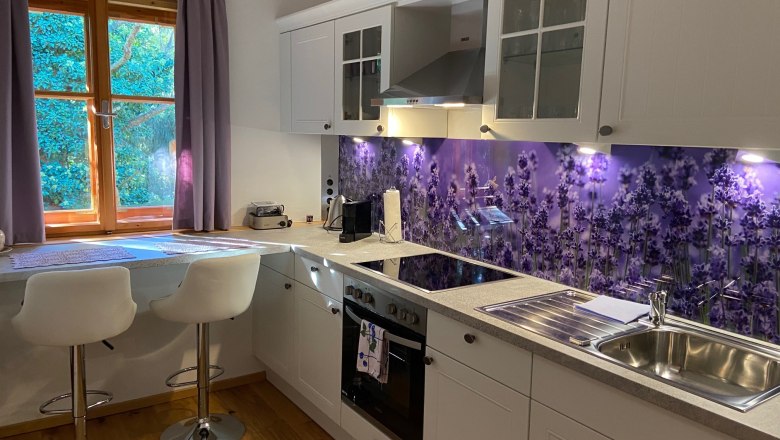 Kitchen, © Wiener Alpen Modern kitchen with white cupboards, lavender splashback and two bar stools in front of a window with purple curtains.