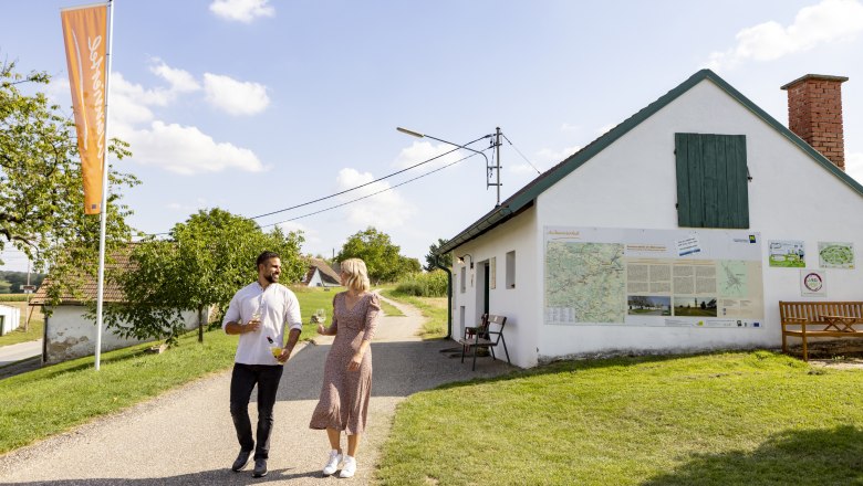 Self-service cellar in the Kettlasbrunn wine cellar lane, © Weinviertel Tourismus GmbH / Schwarz-König A couple walks past a white building with maps on the wall, wine glasses in hand.