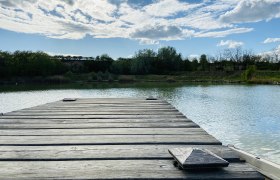 Idyllic fishing paradise, © Weinstraße Weinviertel Wooden footbridge at the brick kiln pond in Zellerndorf, surrounded by trees and a blue sky with clouds.