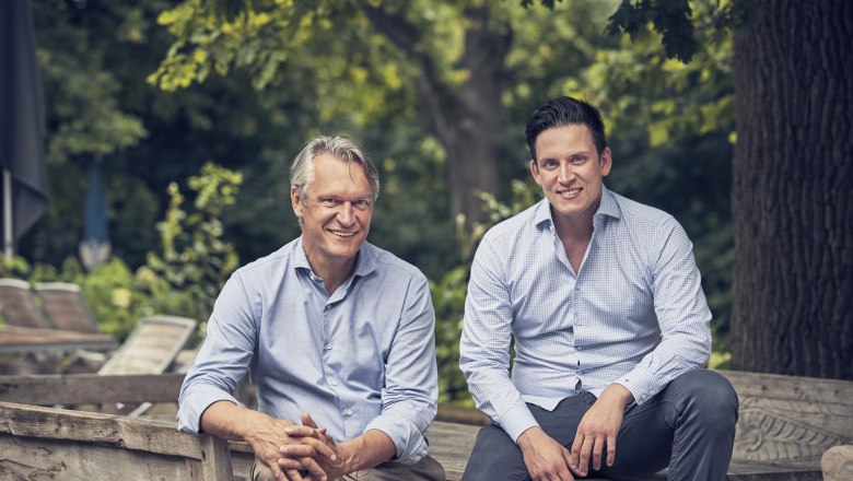 Gerhard Knobl sen. & Gerhard Knobl jun., © Niederösterreich Werbung/Andreas Hofer Two men are sitting on a wooden bench outside, surrounded by trees.