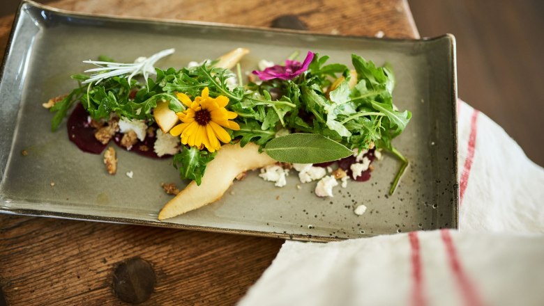 Organic restaurant Leibspeis', © SONNENTOR/ @nudlholz.at An artfully arranged salad with rocket, yellow flower and pear slices on a rectangular plate.