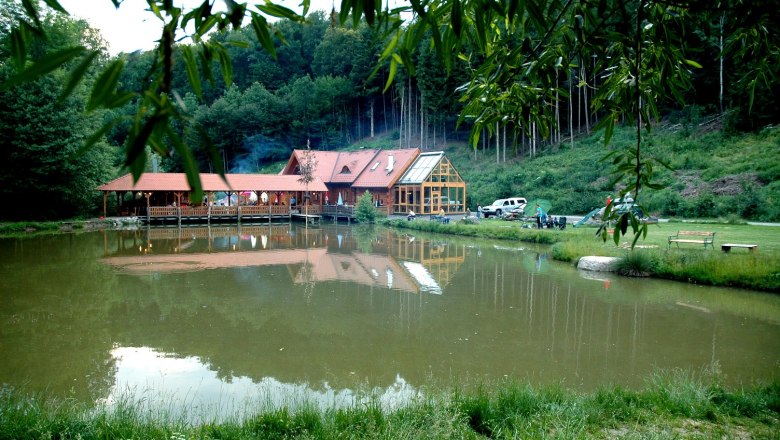 Experience Gaul, © Erlebniswelt Gallien A building with a red roof is reflected in a pond surrounded by trees and meadows.