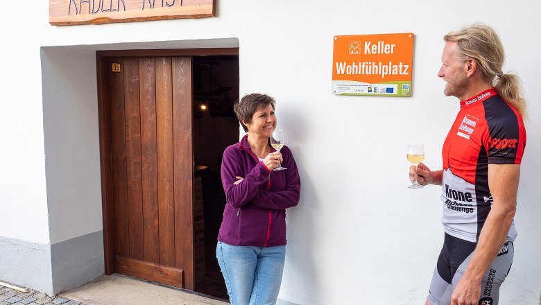 Cellar wellness area Jetzelsdorf, © Initiative Pulkautal / Mödl Two people are standing in front of a building labeled 'Radler-Rast' and 'Keller Wohlfühlplatz', holding glasses of wine.