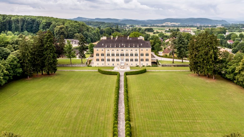 Chateau Petit Versailles, © Château Petit Versailles - David Capellari Aerial view of Frohsdorf Castle, a large, symmetrical castle with a yellow façade, manicured lawn and trees in the background.