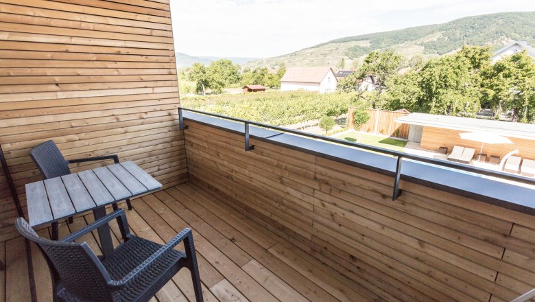 View from the wood-paneled balcony, © KASTNER NEW MEDIA - kastner.tv Wood-paneled balcony with table and two chairs, view of green landscape and hills in the background.