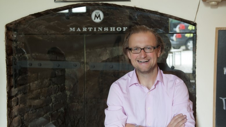 Michael Martin, © Martinshof A man with glasses and a pink shirt stands smiling in front of a sign that reads 'Martinshof'.