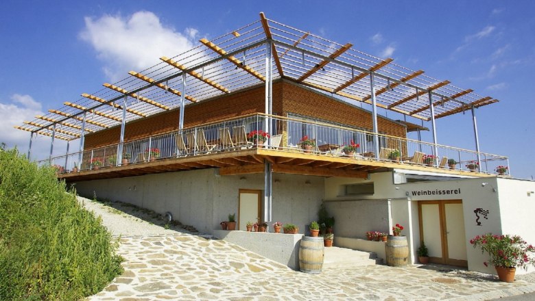 Wine tasting, © Weinbeisserei Hager Modern building with terrace and pergola, surrounded by plants and wine barrels, under a blue sky.