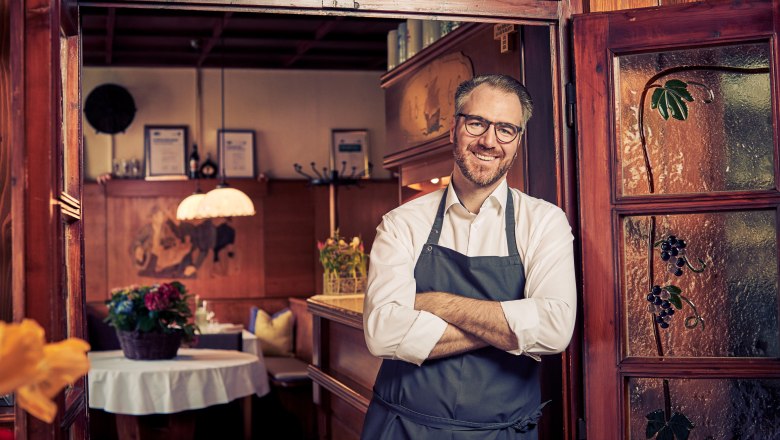 Landlord Thomas Baumgartlinger, © Niederösterreich Werbung/Andreas Hofer Smiling man in an apron stands in the doorway of a cozy restaurant.