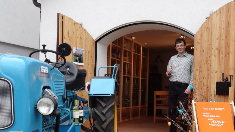 Viticulture Rathammer, © Weinbau Rathammer A man stands with a wine glass in his hand in front of a building with open wooden doors. A blue tractor is parked next to it.