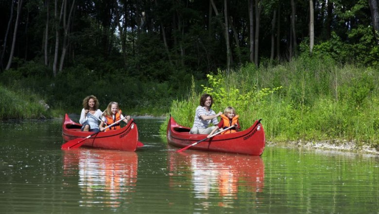 Tulln Water Park, © DIE GARTEN TULLN Two red canoes with adults and children on a calm river, surrounded by green vegetation.