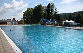Schlossbad Pitten, © Marktgemeinde Pitten A large outdoor pool with clear water, surrounded by trees and hills under a blue sky.