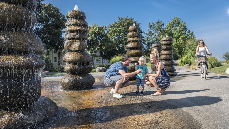 fountain_c_robert-herbst-city-community-tulln, © Robert Herbst/Stadtgemeinde Tulln Family at a fountain in a park, with a woman on a bicycle in the background.