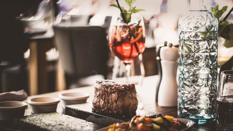 Swiss-style steak, © Route 66 Fehr A juicy steak served on a hot stone, accompanied by vegetables, a carafe of water and a cocktail glass.