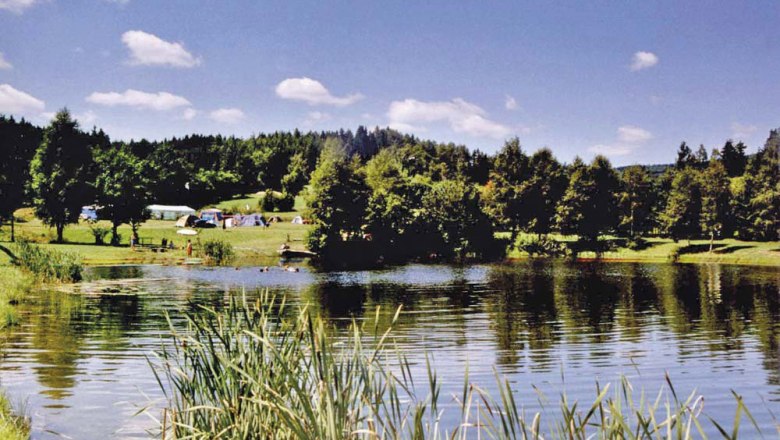 Camping site St. Martin, © Marktgemeinde St. Martin An idyllic campsite with tents on the shore of a lake, surrounded by trees and meadows under a blue sky.
