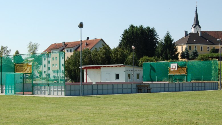Funcourt and youth center, © Gemeinde St. Margarethen A fun court with basketball hoop and green net, with a small building next to it. Residential buildings and a church can be seen in the background.