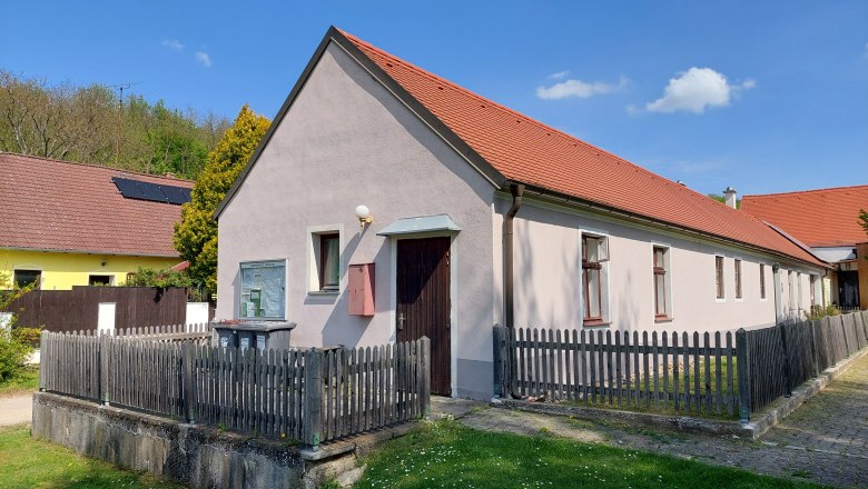 Camping house, © Gemeinde Fallbach A long, single-storey house with a red roof and wooden fence, surrounded by green grass and blue sky.