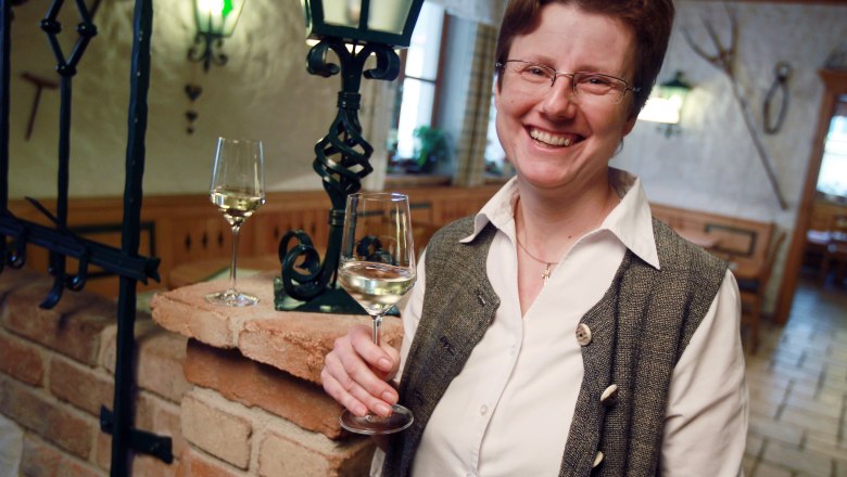 Viticulture Gill, © weinfranz.at A smiling person holds a glass of white wine in a cozy room with a brick wall and a lantern.
