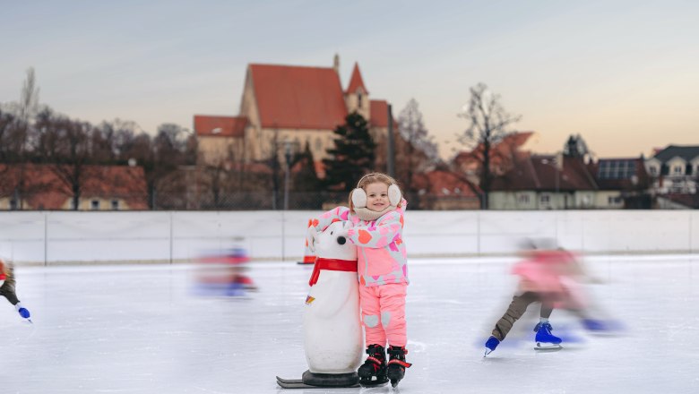 Ice rink in Eggenburg, © Martin Mathes A child in pink clothing stands on an ice rink with a penguin aid. A church can be seen in the background.