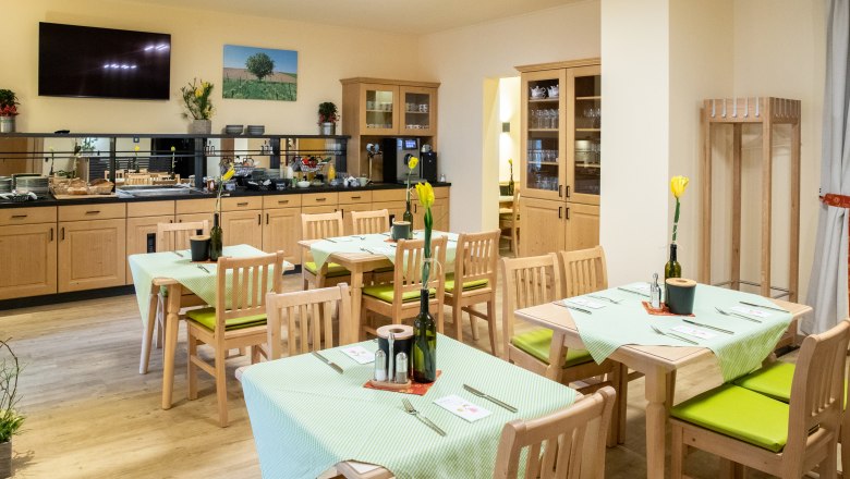 Breakfast room, © Nadine Beles A cozy breakfast room with wooden furniture, laid tables and a buffet area. Pictures hang on the walls and plants decorate the room.