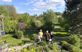 Gitti's garden paradise, © "Natur im Garten" Three people are standing in a green garden with trees and bushes, surrounded by a rural landscape under a blue sky with clouds.
