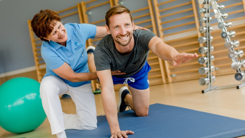 Exercise therapy at the Königsberg health resort, © Niederösterreich Werbung/Doris Schwarz-König A man does an exercise on a mat, supported by a woman in a fitness studio.