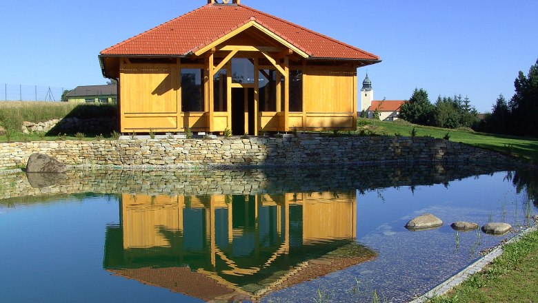 Graduation plant, © Waldenstein A wooden building with a red roof is reflected in a pond, surrounded by a green landscape and blue sky.