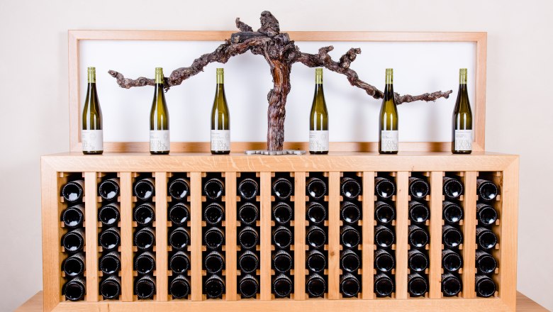 Wine selection at Winzerhof Leonhartsberger, © winzerhof leonhartsberger Wine bottles on a wooden shelf with grapevine decoration.