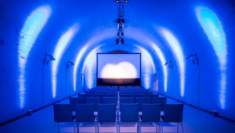 The tube with cinema seating, © Christian Husar A bluish-lit room with cinema seating and a screen.