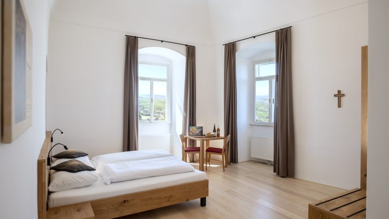 Room example in the guest house at Göttweig Abbey, © Robert Herbst Bright double room with wooden bed, table and chairs, two windows with curtains and a cross on the wall.