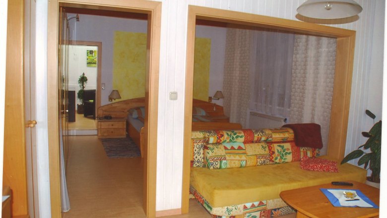 Guest room Weixelbaum, © Weixelbaum A cozy guest room with a colorful sofa, wooden table and a view of a bedroom with a double bed.