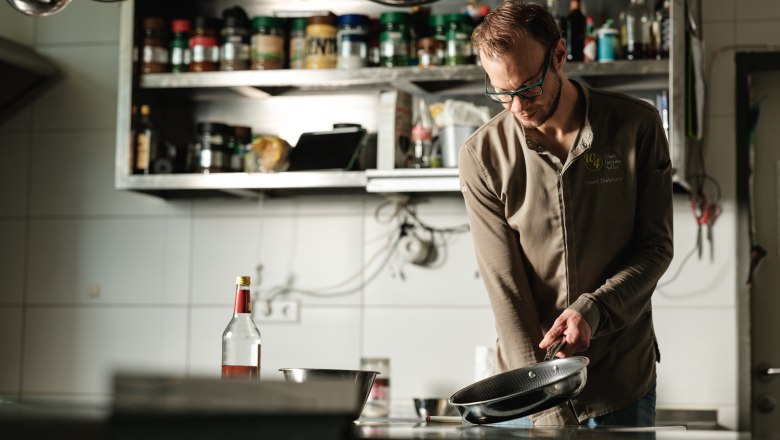 W4, © Weinviertel Tourismus / Michael Reidinger A chef in a professional kitchen holds a pan. Spice racks and kitchen utensils can be seen in the background.