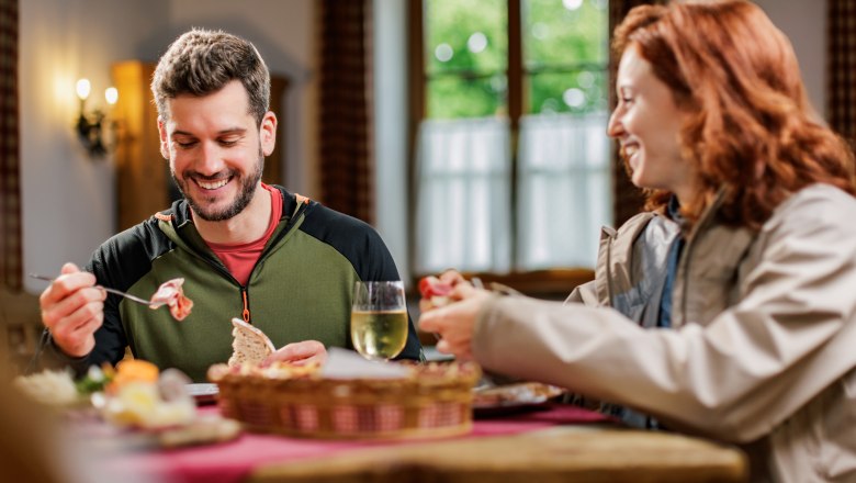 At the wine tavern in Katzelsdorf, © Zwei Wanderer auf einer Holzbrücke im Wald A man and a woman share a snack in a wine tavern