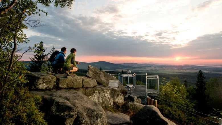 Viewing platform at the Mandelstein, © Studio Kerschbaum Two people on a rock formation at sunset with a view of a viewing platform and vast landscape.