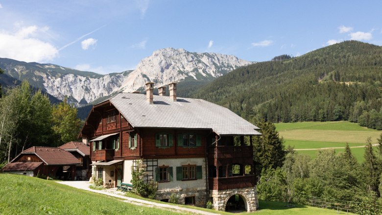 The Riegelhof, © Wiener Alpen - Lierzer A traditional wooden house in a mountainous landscape with meadows and forests in the background.