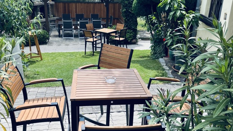 green guest garden, © Anita Gradwohl, dahoam A cozy guest garden with wooden tables and chairs, surrounded by lush greenery and plants.