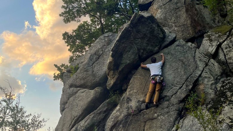 Climbing in Gloxwald, © Familie Moser Person climbing on a rock face outdoors, surrounded by trees and blue sky with clouds.