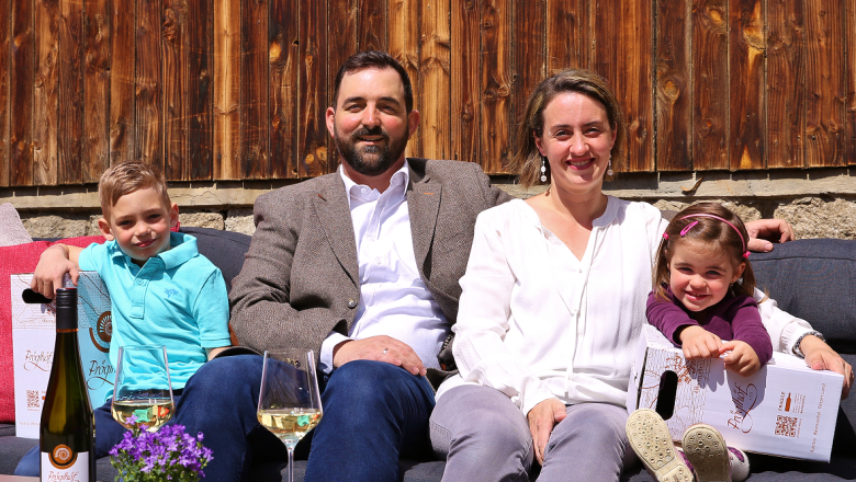 Family Pröglhöf, © Matthias Karasek A family sits on an outdoor sofa in front of a wooden wall, with bottles of wine and glasses on the table.