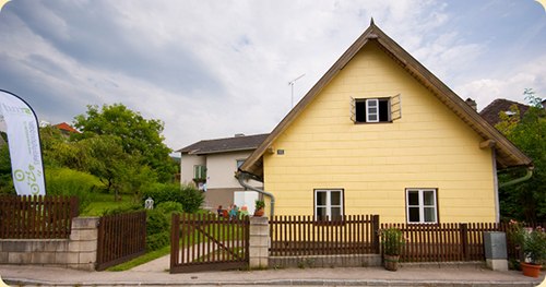 Front view, © srad A yellow house with a pointed roof and wooden fence in a rural setting.