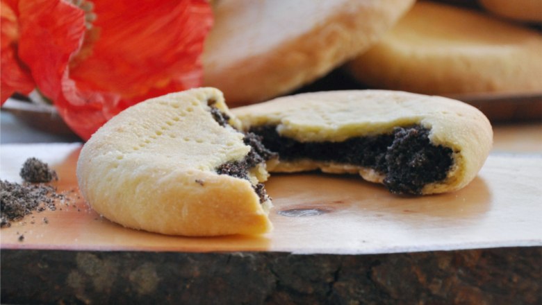 Poppy tents, © Café Pension Kristall A sliced poppy seed tent with poppy seed filling on a wooden board, with a red poppy flower in the background.