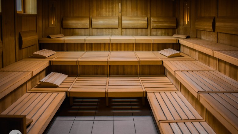 Bio colored light sauna, © TheVerve-Agentur (theverve.at) Interior view of an empty wooden sauna with several rows of seats and headrests.
