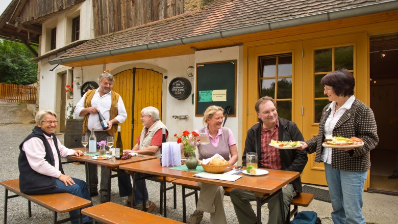 Heurigen snack in front of the house, © Rita Newman Group of people sitting at a table in front of a rustic building enjoying an outdoor meal.