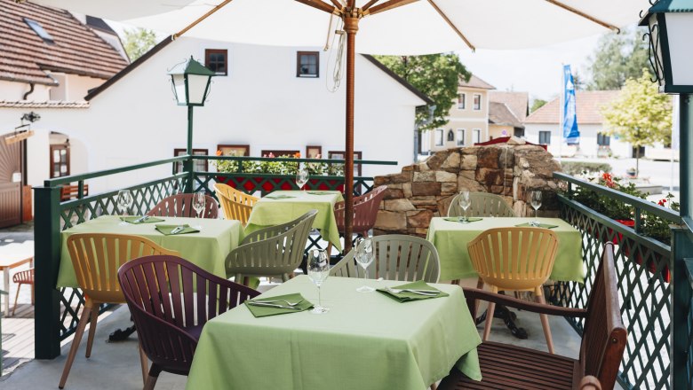 Guest garden located directly on the World Heritage Trail, © Niederösterreich Werbung/David Schreiber A guest garden with green tablecloths and colorful chairs under a parasol, surrounded by buildings and plants.