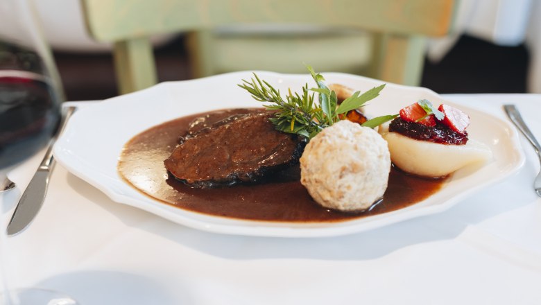 Saddle of venison with bread dumplings, © Niederösterreich Werbung/David Schreiber A plate with saddle of venison, bread dumplings, pear with cranberries and sauce, decorated with herbs.