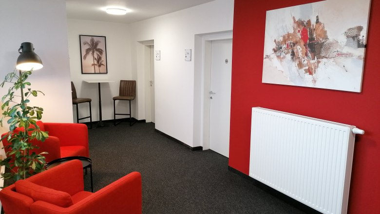 Pension zum Spitz, © Pension zum Spitz Bright hallway with red accents, two red armchairs, a table with chairs and abstract art on the wall.