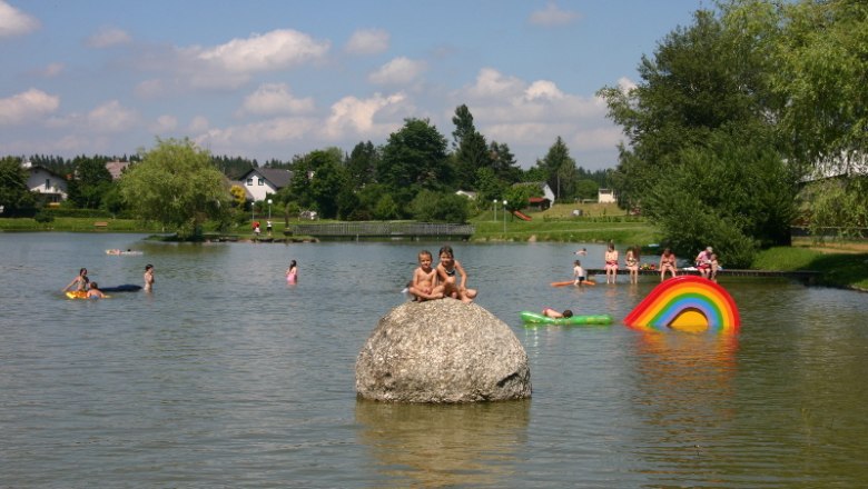 Upper local pond Ottenschlag, © Gemeinde Ottenschlag Children play in the water of a pond with colorful swimming tires and a large stone.