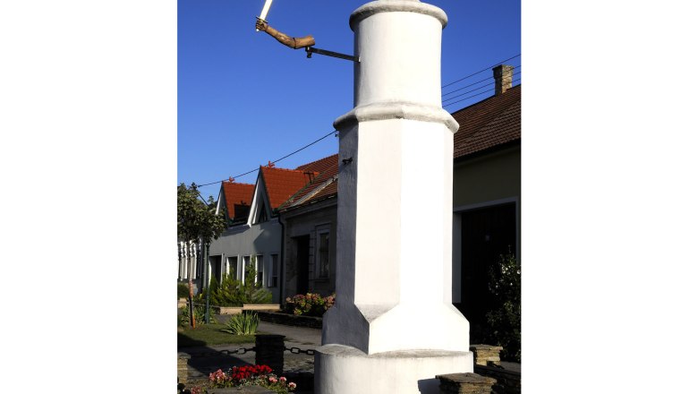 Pillory, © Weinviertel Tourismus / Mandl A white pillory with a wooden hand holding a sword in front of a house with red roofs.