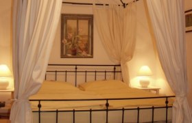 Canopy bed, © Hotel Residenz Schrannenhof A four-poster bed with white curtains in a cozy bedroom, illuminated by two bedside lamps.