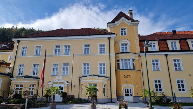Kirchenwirt Hotel, © Karin Stranz Yellow building with red roofs and several windows, surrounded by plants and flags.