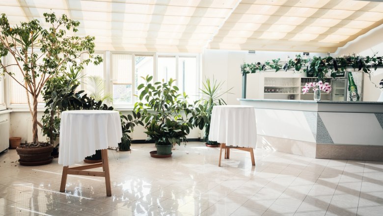 Restaurant, © Auland-Hotel Siebenbrunnerhof Bright room with plants, two bar tables with white tablecloths and a bar in the background.