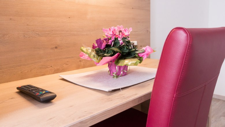 Double room, © W. Garhöfer A desk with a remote control, a bouquet of flowers and a red chair.